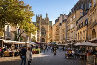 A panoramic view of Metz city center with the cathedral, golden stone buildings, busy pedestrian streets, and café terraces in warm sunlight.